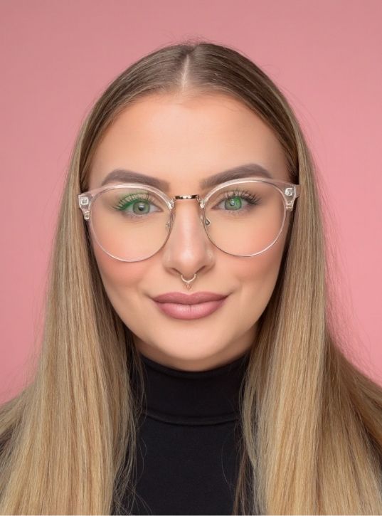 A woman with long straight blonde hair, wearing clear glasses, a black top, and a septum piercing, smiles slightly against a pink background.