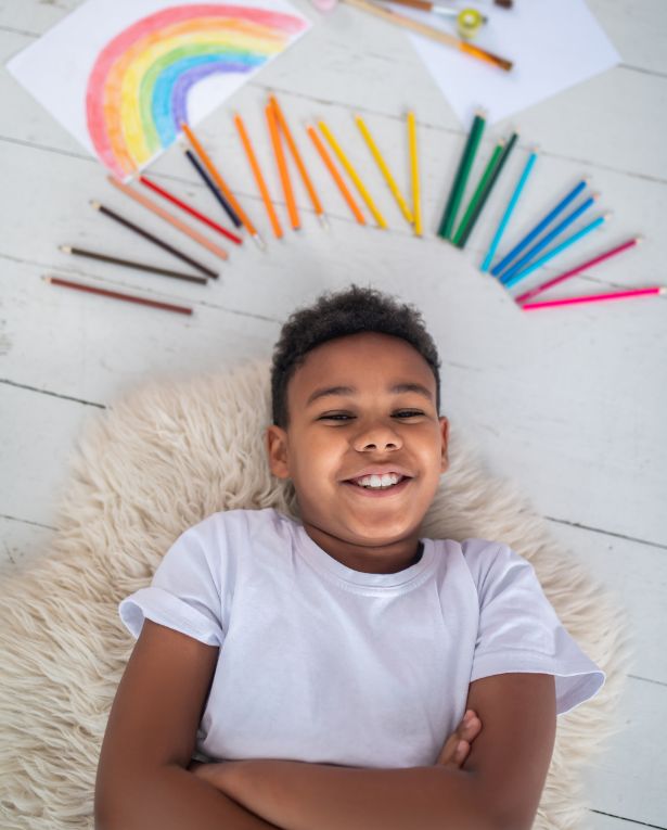 A smiling boy lies on a fluffy rug with his arms crossed. Above his head are colored pencils arranged in a semi-circle and a colorful rainbow drawing on white paper. The scene is bright and cheerful.