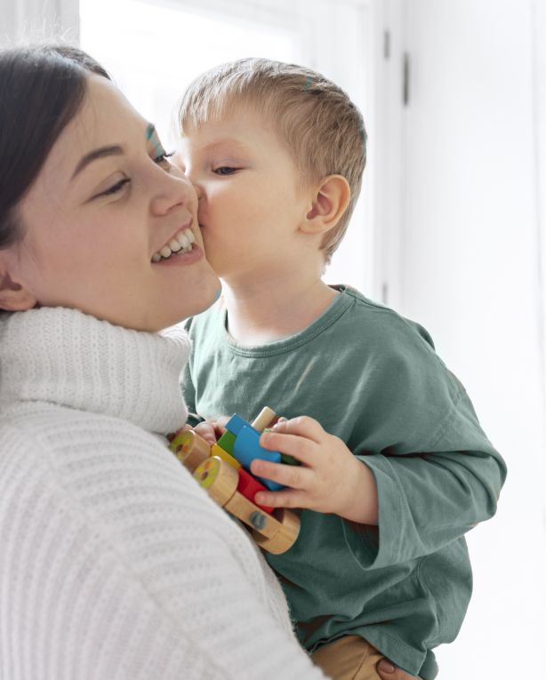 A young child kisses a smiling woman on the cheek while holding colorful wooden toys. The woman wears a white turtleneck sweater and the child is dressed in a green shirt. They are indoors in a brightly lit room.