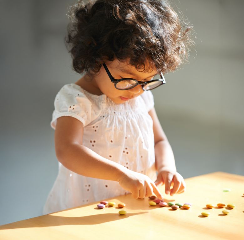 A young child with curly hair and glasses is sorting colorful candy or beads on a wooden table, focused on the activity in natural light.
